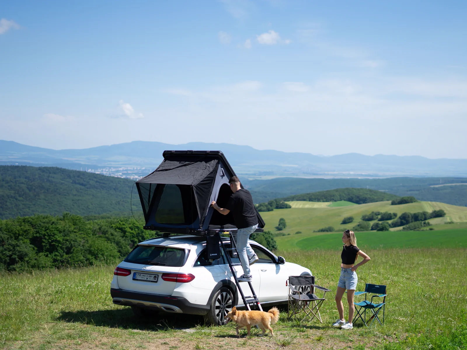 Couple installant une tente de toit SNAILER sur une voiture dans une prairie de montagne